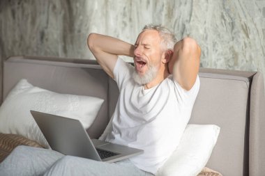 Laptopla gözleri kapalı esneyen bir adam.