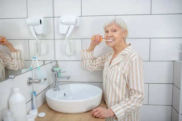 Pijamalı yaşlı bir kadın banyoda dişlerini fırçalıyor. — Stok fotoğraf