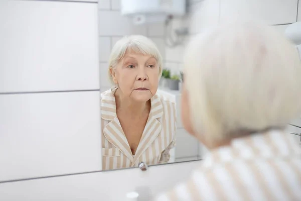 Femme aux cheveux gris regardant dans le miroir dans la salle de bain — Photo