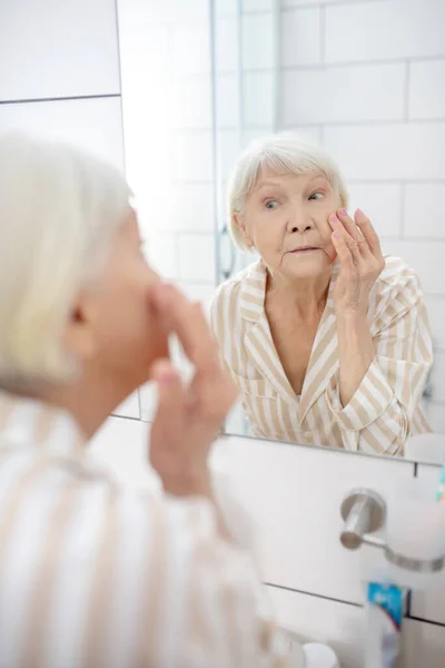 Grauhaarige Frau blickt im Badezimmer in den Spiegel und berührt ihr Gesicht — Stockfoto