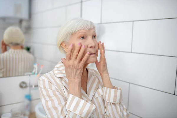 Femme aux cheveux gris regardant dans le miroir dans la salle de bain et touchant ses joues — Photo