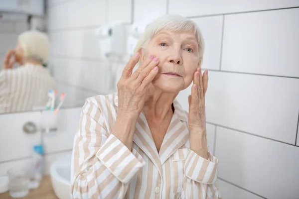 Femme aux cheveux gris regardant dans le miroir dans la salle de bain et touchant son visage — Photo