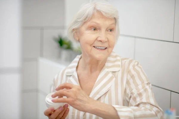 Mujer de pelo gris sosteniendo un frasco de crema y mirando a un lado — Foto de Stock