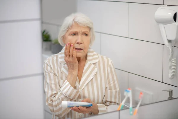 Grijs harige vrouw in pyjama loking op zichzelf in de spiegel en het gevoel gefrustreerd — Stockfoto