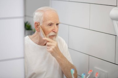 Beyaz tişörtlü, sakalını fırçalayan gri saçlı adam.