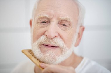 Beyaz tişörtlü yaşlı adam sakalını fırçalıyor.