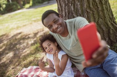 Koyu tenli gülümseyen adam parkta kızıyla selfie çekiyor.