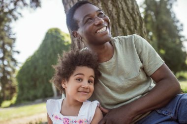 Koyu tenli bir adam çocuğunun yanında oturuyor ve gülümsüyor.