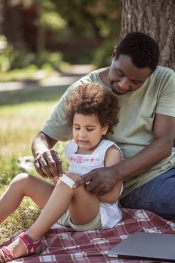 Koyu tenli bir adam, kızının dizine yara bandı yapıştırıyor.