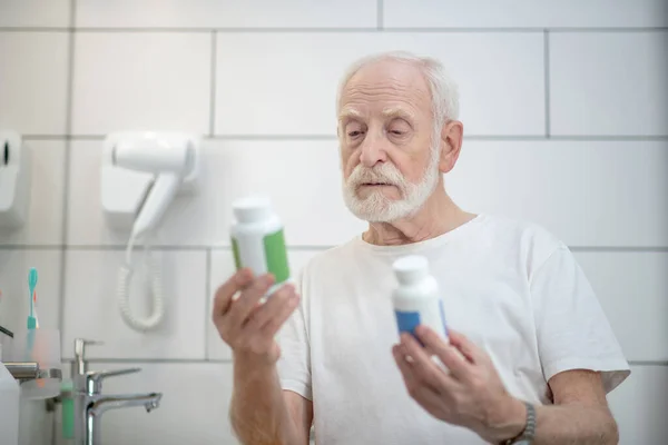 Gray-haired man in white tshirt holding bottles with pills and looking thoughtful — Stock Photo, Image