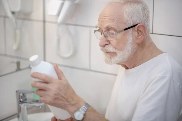 Сіроволосий чоловік в окулярах вивчає пляшку шампуню — стокове фото