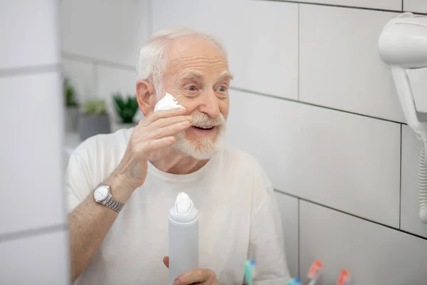 Hombre de pelo gris en camiseta blanca poniendo espuma de afeitar en su cara —  Fotos de Stock