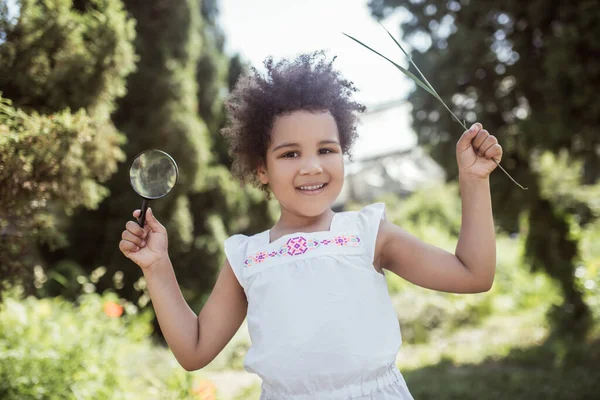 Kudrnatá holčička drží lupu a vypadá vesele — Stock fotografie