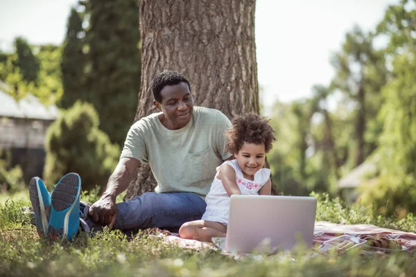Krullend klein meisje zitten onder de boom met haar vader en kijken naar iets online — Stockfoto