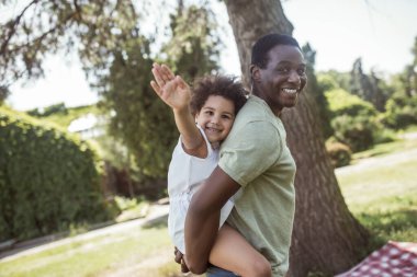 Şirin kıvırcık çocuk babasının sırtında oturuyor ve gülümsüyor.