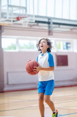 Siyah saçlı spor giysili çocuk topu basketbol sahasına atmaya hazır.