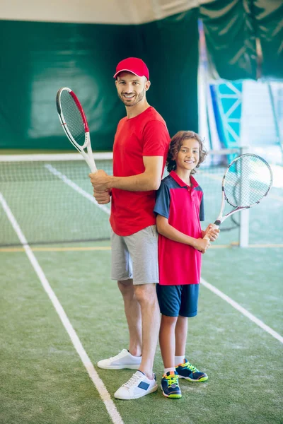 Trener w czerwonej czapce i chłopiec trzymający rakiety tenisowe w rękach — Zdjęcie stockowe
