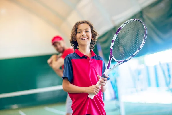 Roztomilý tmavovlasý chlapec stojící s tenisovou raketou a s úsměvem — Stock fotografie