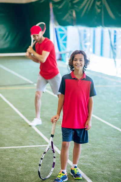 Bonito menino de cabelos escuros de pé com uma raquete de tênis e sorrindo — Fotografia de Stock