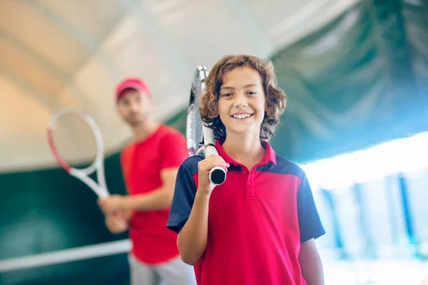 Donkerharige jongen staand met een tennisracket en glimlachend — Stockfoto