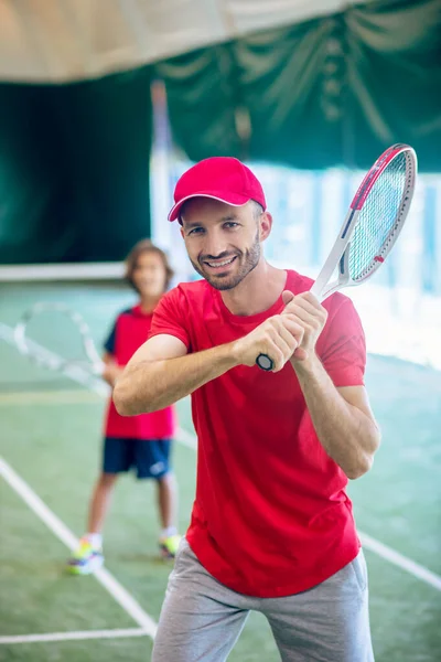 Молодой бородатый мужчина в красной кепке машет теннисной ракеткой — стоковое фото