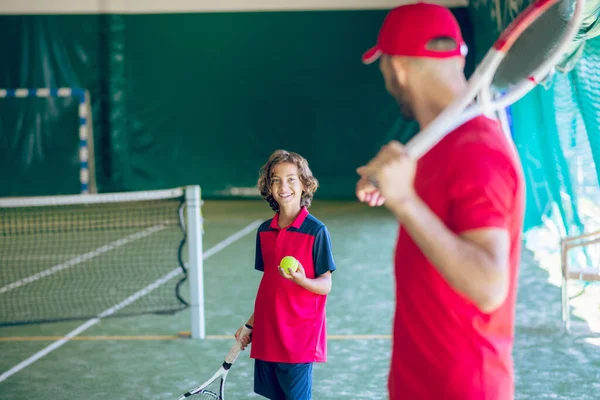 Jovem barbudo com um boné vermelho segurando uma raquete de tênis e falando com um menino — Fotografia de Stock