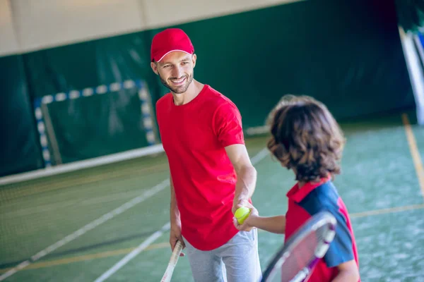 Homem barbudo jovem em um boné vermelho apertando as mãos com um menino de cabelos escuros antes de jogar tênis — Fotografia de Stock