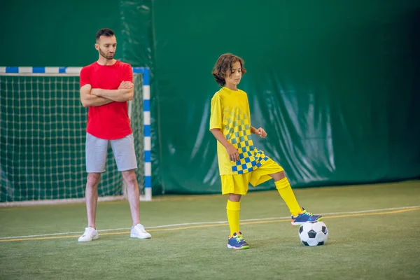 Lindo chico en uniforme amarillo jugando fútbol, su entrenador mirándolo — Foto de Stock