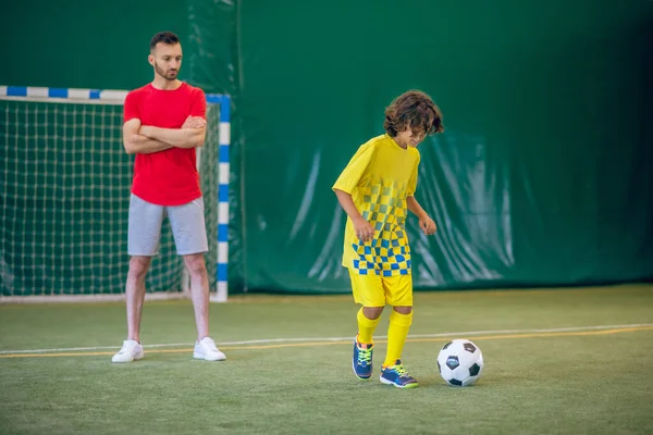 Lindo chico en uniforme amarillo jugando fútbol, su entrenador mirándolo — Foto de Stock