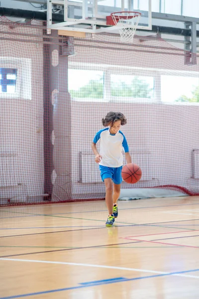 Menino de cabelos escuros em sportswear correndo atrás da bola em um ginásio — Fotografia de Stock
