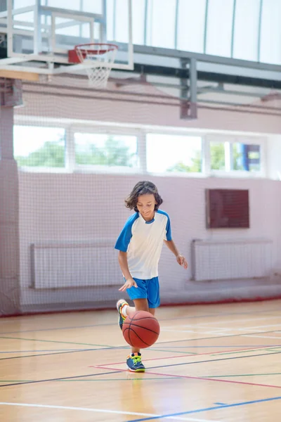Menino de cabelos escuros em sportswear correndo atrás da bola em um ginásio — Fotografia de Stock