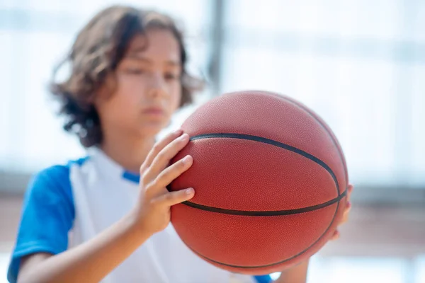 Menino de cabelos escuros em sportswear segurando uma bola e olhando sério — Fotografia de Stock