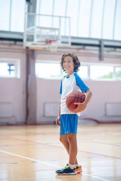 Menino de cabelos escuros em sportswear em pé no ginásio com um bal nas mãos — Fotografia de Stock