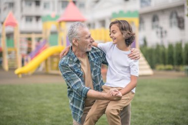 Gri saçlı adam oğluna sarılıyor ve mutlu görünüyor.