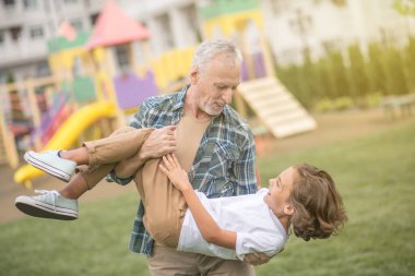 Gri saçlı adam oğluyla oynuyor ve mutlu görünüyor.