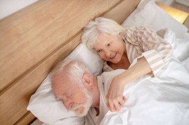 Gri saçlı yaşlı kadın uyuyan kocasına sarılıyor.