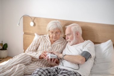 Gri saçlı yaşlı adam, karısıyla birlikte tansiyonunu ölçüyor.