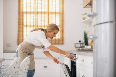 Önlüklü kadın mutfakta fırını açıyor.