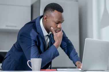 Koyu tenli zarif bir adam dizüstü bilgisayardan bir şey okuyor ve düşünceli görünüyor.