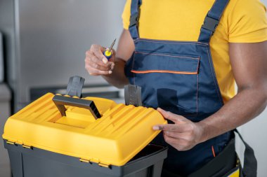 Koyu tenli servis elemanı mutfakta duruyor ve elinde aletlerle bir kutu tutuyor.