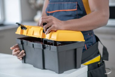 Koyu tenli servis elemanı mutfakta duruyor ve alet kutusunu açıyor.