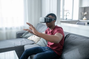 Siyah tenli genç bir adam VR gözlükleriyle oturuyor ve eğleniyor.