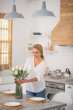 Kot pantolonlu sarışın ev kadını vazoya çiçek koyuyor.