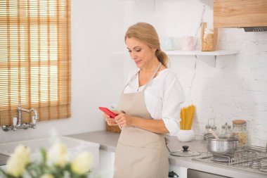 Önlüklü kadın elinde tabletle mutfakta duruyor.