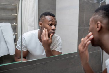 Koyu tenli genç yakışıklı adam aynada kendine bakıyor.