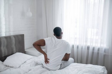 Afro-Amerikalı bir adam yatakta oturuyor ve sırtına dokunuyor.