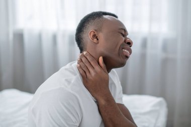 Afro-Amerikalı bir adam yatakta oturup boynuna dokunuyor.