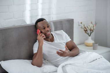 Beyaz tişörtlü genç bir adam yatakta uzanıyor ve telefonda konuşuyor.
