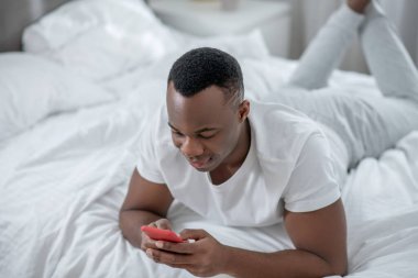 Beyaz tişörtlü genç bir adam yatakta uzanıyor ve internette sohbet ediyor.