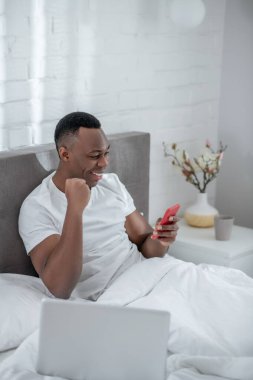 Beyaz tişörtlü genç bir adam yatakta uzanıyor ve uzaktan çalışıyor.